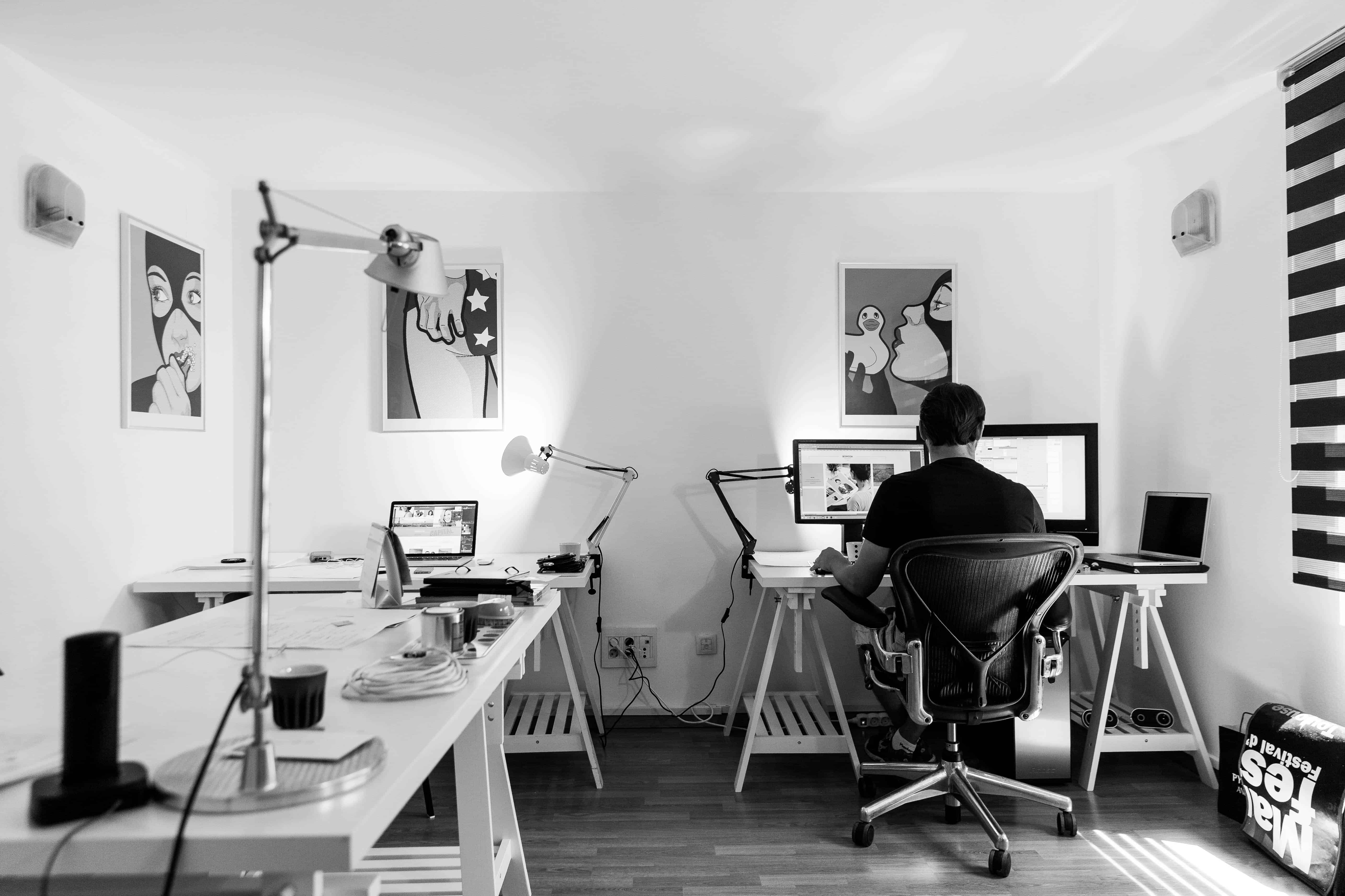 Black and white photo of a person working at a computer desk in a minimal home office with artwork on the walls