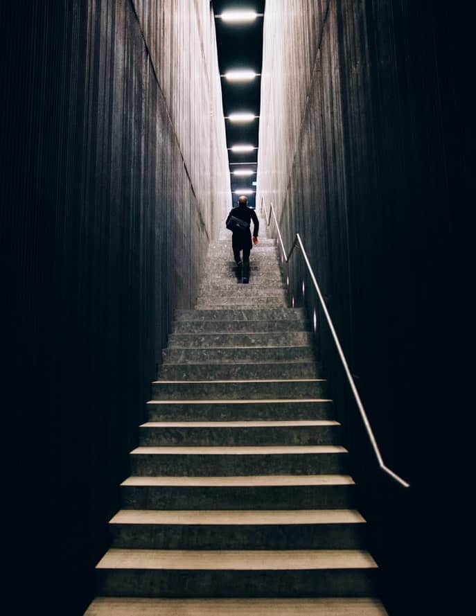Silhouette of a person climbing a dramatic staircase between tall dark walls toward a lit ceiling