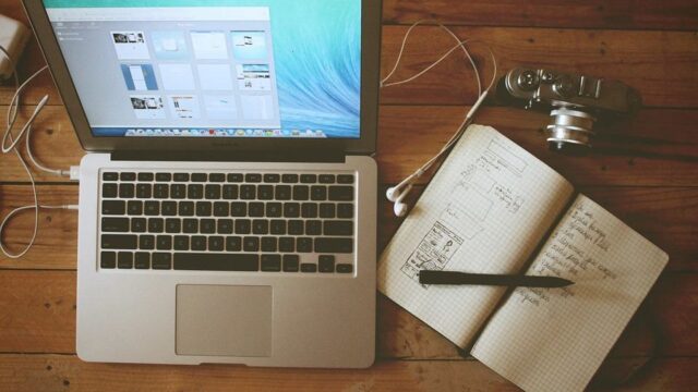 MacBook, open notebook with wireframe sketches, earphones, and a vintage camera on a wooden desk